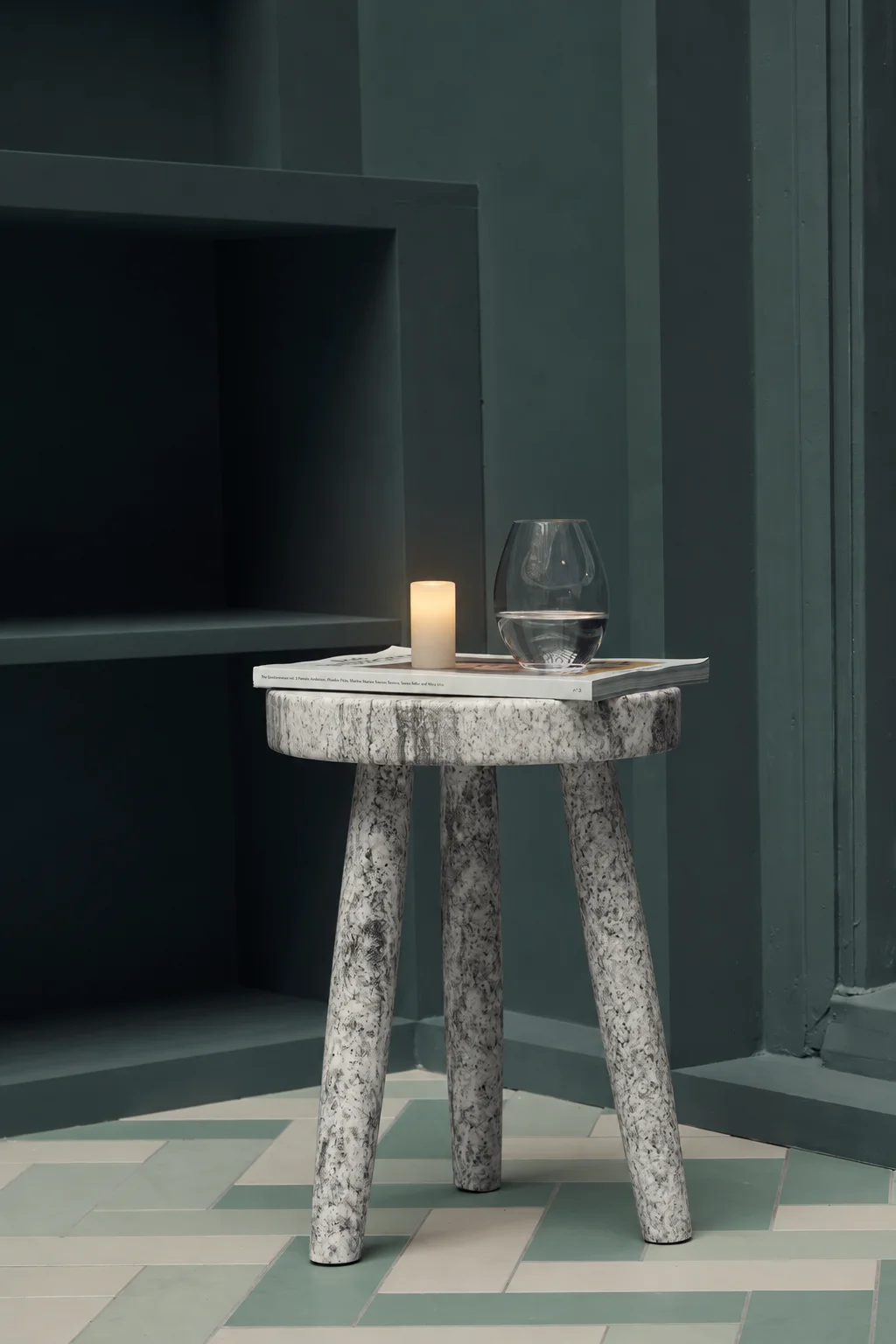 A Context candle and glass on a sculptural stone side table against dark wall panels
